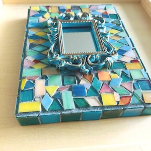 May include: A rectangular mosaic frame featuring a central, ornate, metallic blue mirror with a decorative border. The surrounding surface is covered in a dense arrangement of multi-colored glass tiles in shades of blue, green, yellow, and pink.