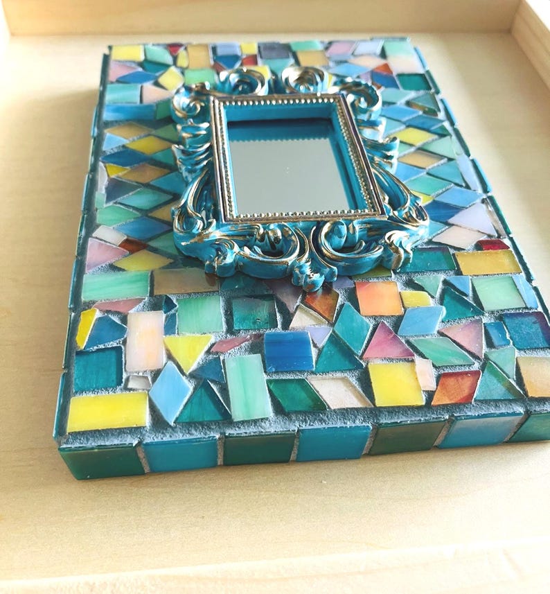 May include: A rectangular mosaic frame featuring a central, ornate, metallic blue mirror with a decorative border. The surrounding surface is covered in a dense arrangement of multi-colored glass tiles in shades of blue, green, yellow, and pink.