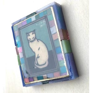 May include: A rectangular decorative box featuring a central illustration of a white cat on a dark background with the text A.J.D.G 1917. The frame is composed of small, multi-colored glass tiles in shades of blue, pink, green, and purple.