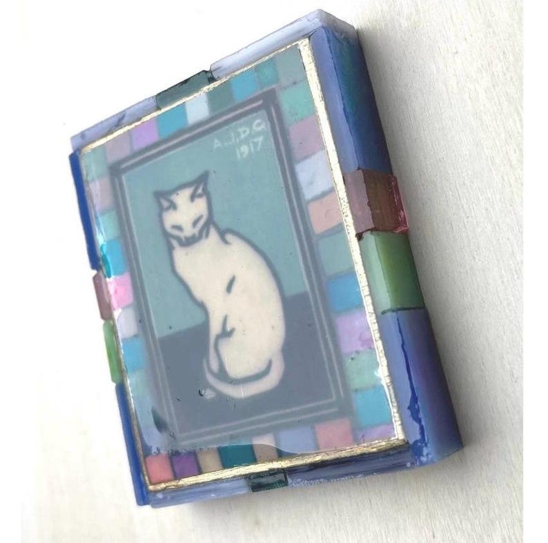 May include: A rectangular decorative box featuring a central illustration of a white cat on a dark background with the text A.J.D.G 1917. The frame is composed of small, multi-colored glass tiles in shades of blue, pink, green, and purple.