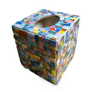 May include: A square tissue box cover decorated with a multi-colored mosaic pattern. The exterior features small, irregularly shaped glass tiles in shades of blue, yellow, pink, orange, and green, set against a light-colored grout base.