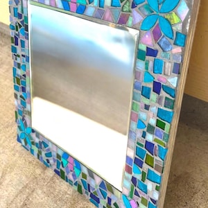 Puede incluir: Un espejo cuadrado con un marco de mosaico decorativo compuesto por varias piezas de vidrio en tonos azul, verde, morado y blanco iridiscente. El marco incluye dos arreglos de piezas en forma de flor fijados con lechada gris.