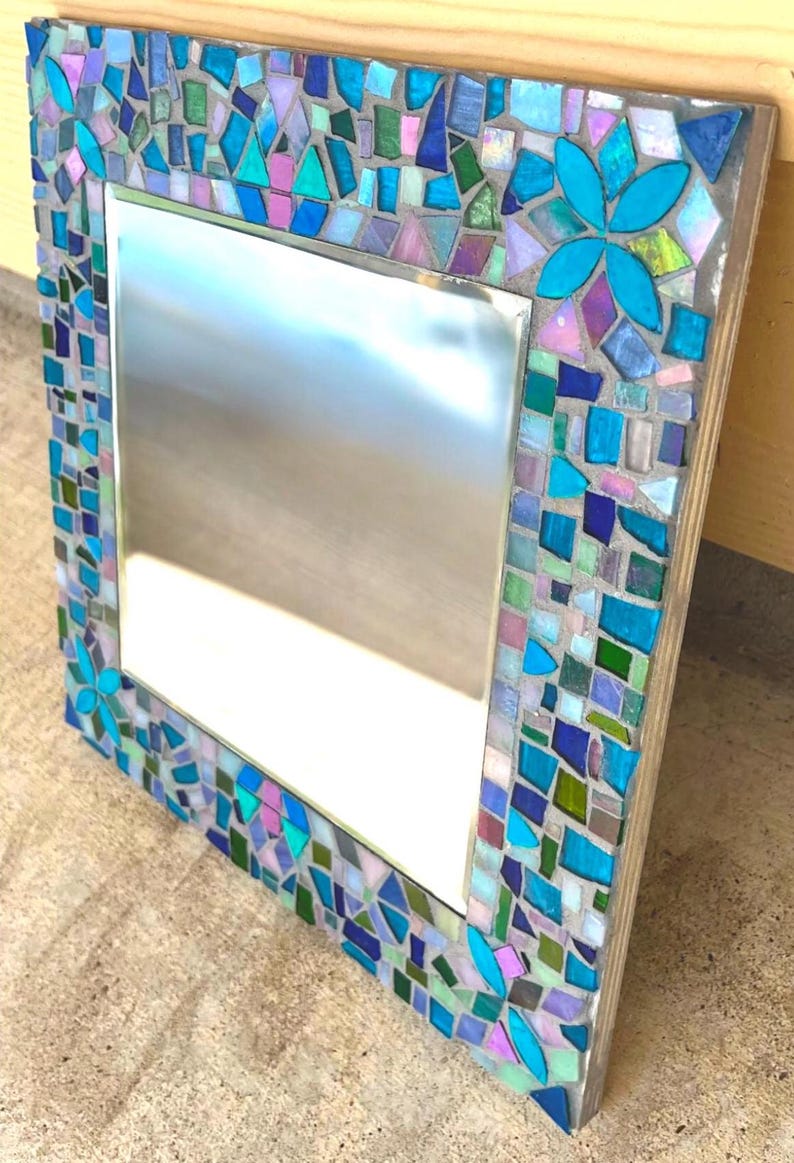 Puede incluir: Un espejo cuadrado con un marco de mosaico decorativo compuesto por varias piezas de vidrio en tonos azul, verde, morado y blanco iridiscente. El marco incluye dos arreglos de piezas en forma de flor fijados con lechada gris.