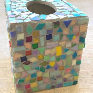 May include: A square tissue box cover featuring a mosaic design made of small, multi-colored glass tiles in shades of blue, green, yellow, pink, and white, all set into a light gray grout base with a circular opening at the top for dispensing.