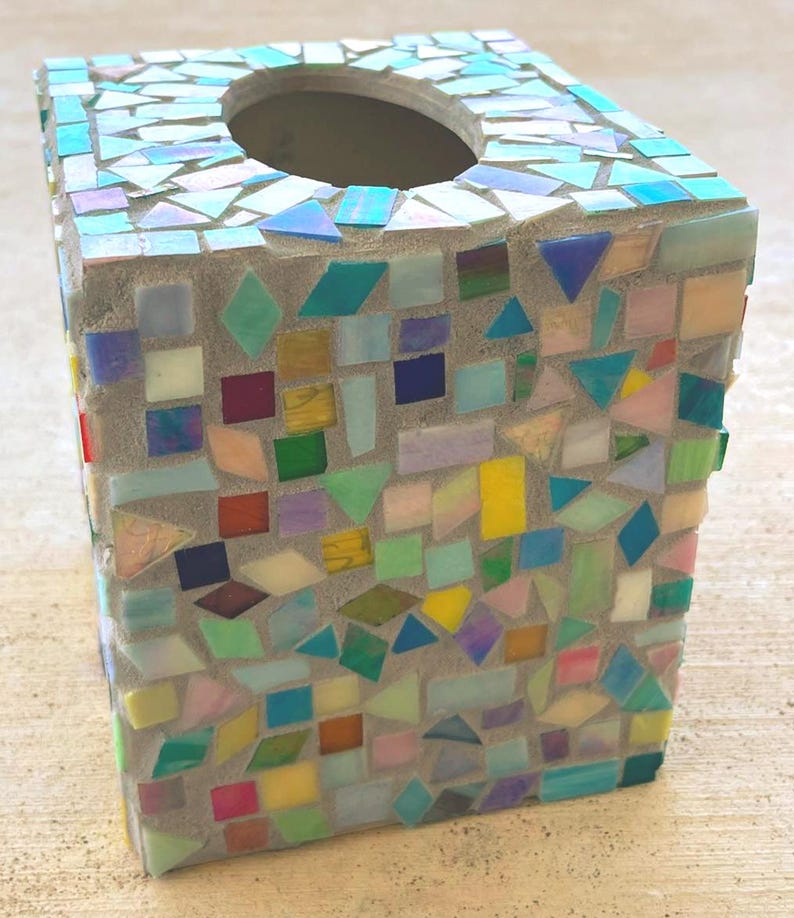 May include: A square tissue box cover featuring a mosaic design made of small, multi-colored glass tiles in shades of blue, green, yellow, pink, and white, all set into a light gray grout base with a circular opening at the top for dispensing.