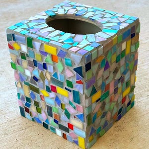May include: A square tissue box cover decorated with a mosaic of multicolored glass tiles in shades of blue, green, yellow, red, and pink. The tiles are set in light gray grout, creating a textured surface with an oval opening on the top side.