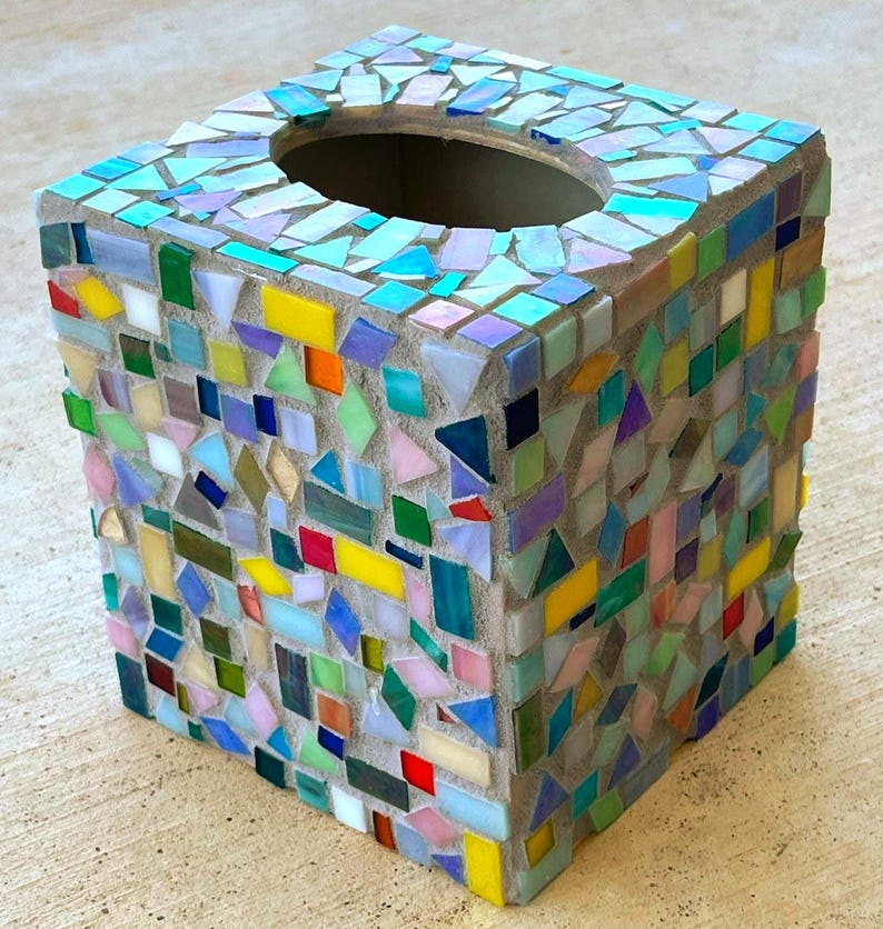 May include: A square tissue box cover decorated with a mosaic of multicolored glass tiles in shades of blue, green, yellow, red, and pink. The tiles are set in light gray grout, creating a textured surface with an oval opening on the top side.