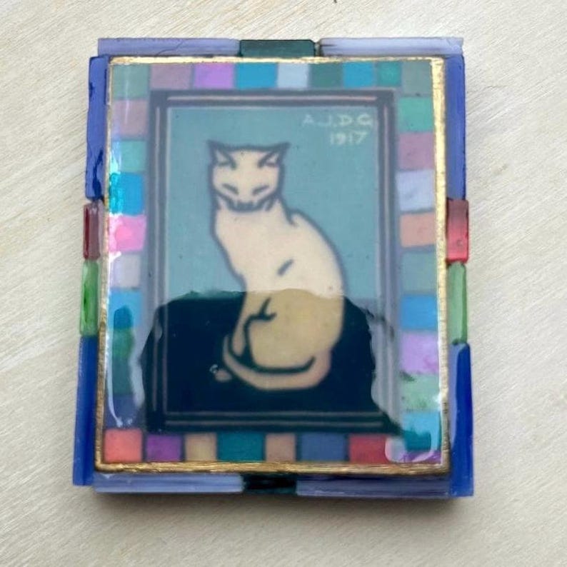 May include: A rectangular decorative tile featuring a central illustration of a seated cream-colored cat on a dark blue background. The image includes the text A.J.D.G 1917. The center is framed by a border of small, multicolored glass mosaic tiles.