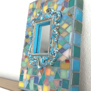 May include: A rectangular mirror featuring a frame decorated with a mosaic of multicolored glass tiles in shades of blue, yellow, green, and orange. Centered on the mosaic is a smaller, ornate, metallic blue and gold-toned rectangular frame.