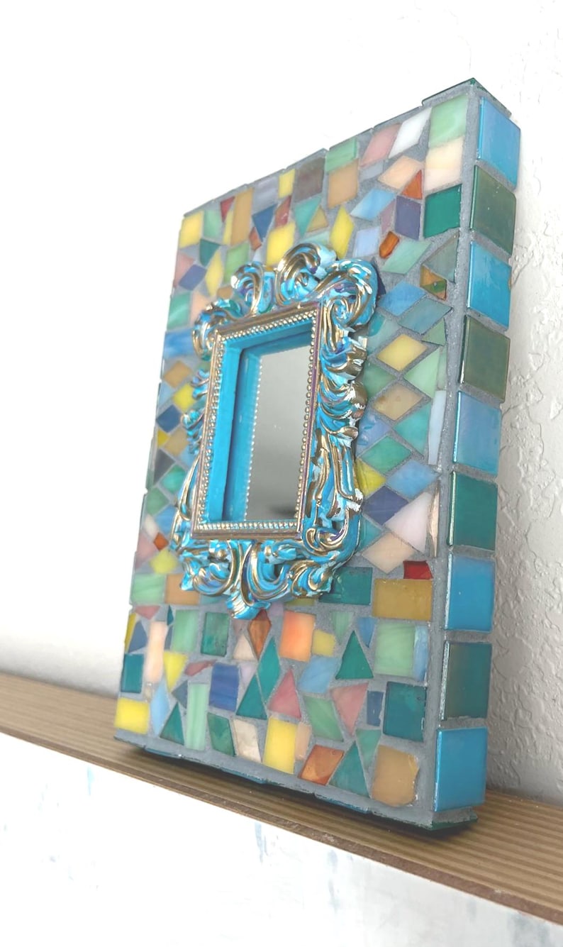 May include: A rectangular mirror featuring a frame decorated with a mosaic of multicolored glass tiles in shades of blue, yellow, green, and orange. Centered on the mosaic is a smaller, ornate, metallic blue and gold-toned rectangular frame.
