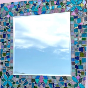 Puede incluir: Un espejo de pared cuadrado con un marco de mosaico decorativo compuesto por varios azulejos de vidrio en tonos turquesa, azul, verde y morado iridiscente. El marco incluye patrones florales repetitivos en las esquinas y los bordes.