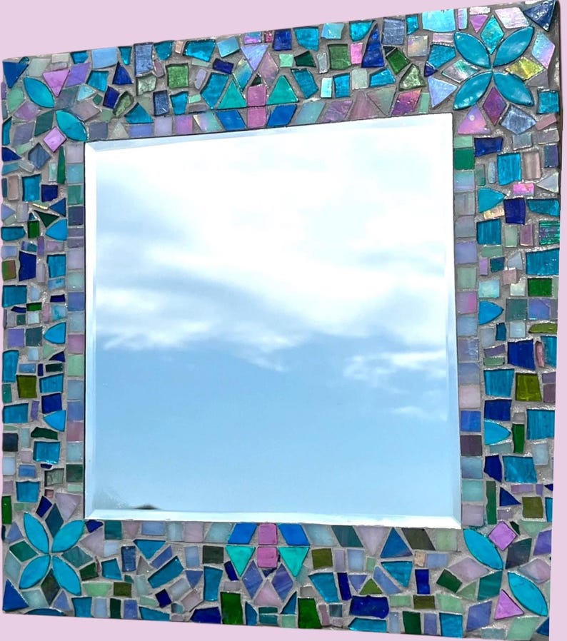 Puede incluir: Un espejo de pared cuadrado con un marco de mosaico decorativo compuesto por varios azulejos de vidrio en tonos turquesa, azul, verde y morado iridiscente. El marco incluye patrones florales repetitivos en las esquinas y los bordes.