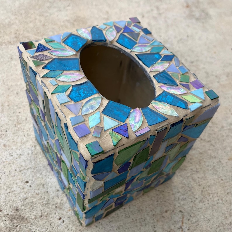 Op de afbeelding: Een vierkante tissueboxhouder versierd met een moza&iuml;ek van glasstukjes in diverse tinten blauw, groen en iriserend wit. De tegeltjes zijn in een willekeurig patroon geplaatst en ingevoegd met lichtgekleurde mortel, met een ovale opening aan de bovenzijde.