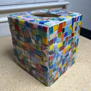 May include: A square tissue box cover featuring a multi-colored mosaic design. The exterior is covered in small, irregularly shaped glass tiles in shades of red, orange, yellow, green, blue, and pink, all set against a white grout base.