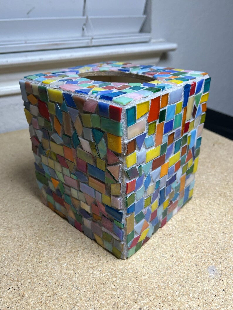 May include: A square tissue box cover featuring a multi-colored mosaic design. The exterior is covered in small, irregularly shaped glass tiles in shades of red, orange, yellow, green, blue, and pink, all set against a white grout base.
