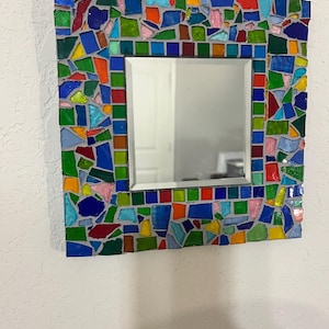 May include: A square wall mirror featuring a decorative frame composed of multi-colored glass mosaic tiles in shades of blue, green, red, yellow, and pink, set in gray grout. The central mirror has a beveled edge and a reflective surface.