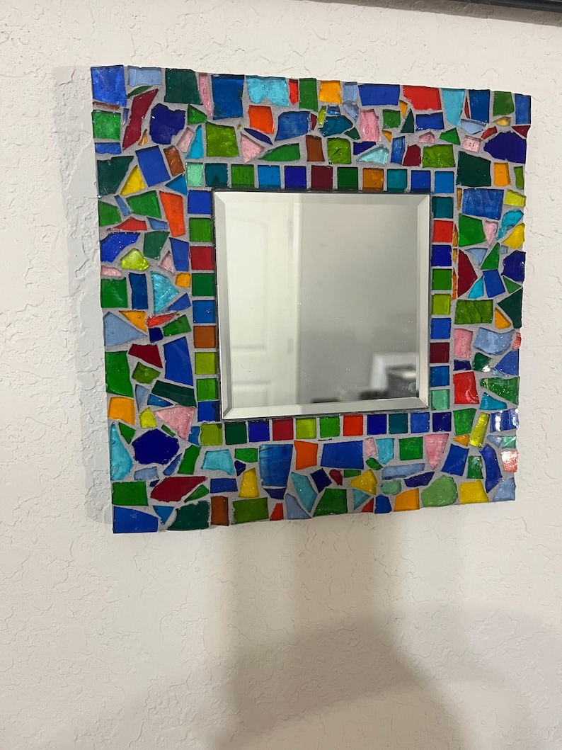 May include: A square wall mirror featuring a decorative frame composed of multi-colored glass mosaic tiles in shades of blue, green, red, yellow, and pink, set in gray grout. The central mirror has a beveled edge and a reflective surface.
