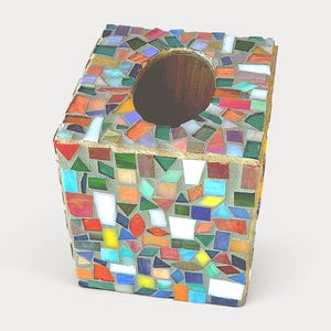 May include: A square tissue box cover featuring a mosaic design made of small, multi-coloured glass pieces in shades of red, blue, green, yellow, and white, set in grey grout with a circular opening at the top and gold-toned edges.