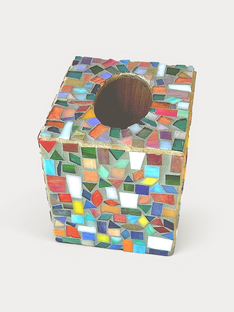 May include: A square tissue box cover featuring a mosaic design made of small, multi-coloured glass pieces in shades of red, blue, green, yellow, and white, set in grey grout with a circular opening at the top and gold-toned edges.