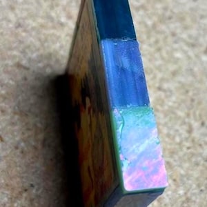 May include: A rectangular block with a layered side profile featuring a dark blue top section, a translucent light blue middle section, and a vibrant iridescent green and pink bottom section, attached to a flat panel with a warm-toned graphic.