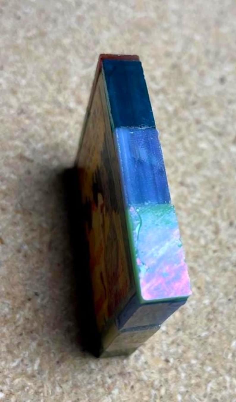 May include: A rectangular block with a layered side profile featuring a dark blue top section, a translucent light blue middle section, and a vibrant iridescent green and pink bottom section, attached to a flat panel with a warm-toned graphic.