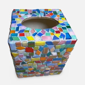 May include: A square tissue box cover decorated with a mosaic of small, multi-colored glass tiles in shades of blue, green, yellow, orange, red, and pink. The tiles are set in white grout, and the top features a large, centered oval opening.