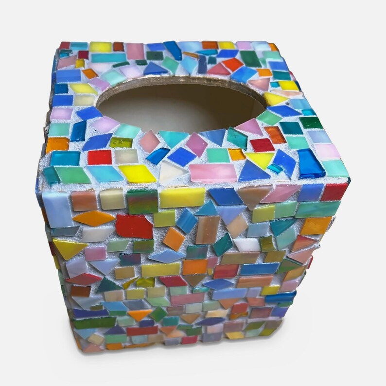 May include: A square tissue box cover decorated with a mosaic of small, multi-colored glass tiles in shades of blue, green, yellow, orange, red, and pink. The tiles are set in white grout, and the top features a large, centered oval opening.