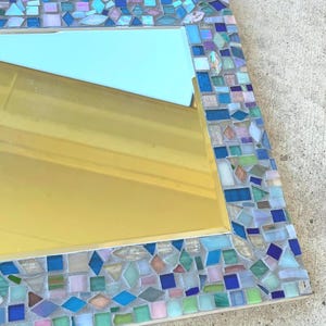 Op de afbeelding: Een rechthoekige wandspiegel met een decoratieve moza&iuml;eklijst bestaande uit kleine, onregelmatig gevormde glazen tegels in tinten blauw, groen, roze en wit. Het reflecterende oppervlak biedt een helder zicht op de omgeving.