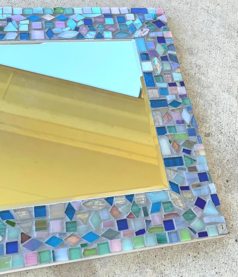Op de afbeelding: Een rechthoekige wandspiegel met een decoratieve moza&iuml;eklijst bestaande uit kleine, onregelmatig gevormde glazen tegels in tinten blauw, groen, roze en wit. Het reflecterende oppervlak biedt een helder zicht op de omgeving.