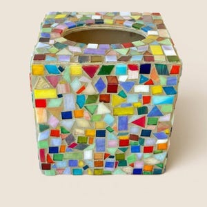 May include: A square tissue box cover featuring a multi-colored mosaic design made of small, irregular glass tiles in shades of red, blue, yellow, green, and white, set into a light-colored grout with a circular opening at the top center.
