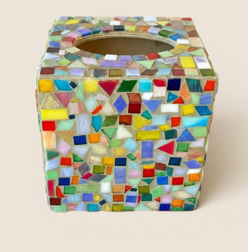 May include: A square tissue box cover featuring a multi-colored mosaic design made of small, irregular glass tiles in shades of red, blue, yellow, green, and white, set into a light-colored grout with a circular opening at the top center.