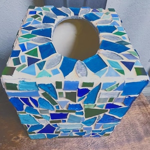 Op de afbeelding: Een vierkante tissueboxhouder met een moza&iuml;ekontwerp van diverse tinten blauw, groen en helder glas, ingebed in lichtgekleurde voegspecie. De bovenzijde is voorzien van een centrale ovale opening voor het uitgeven van tissues.