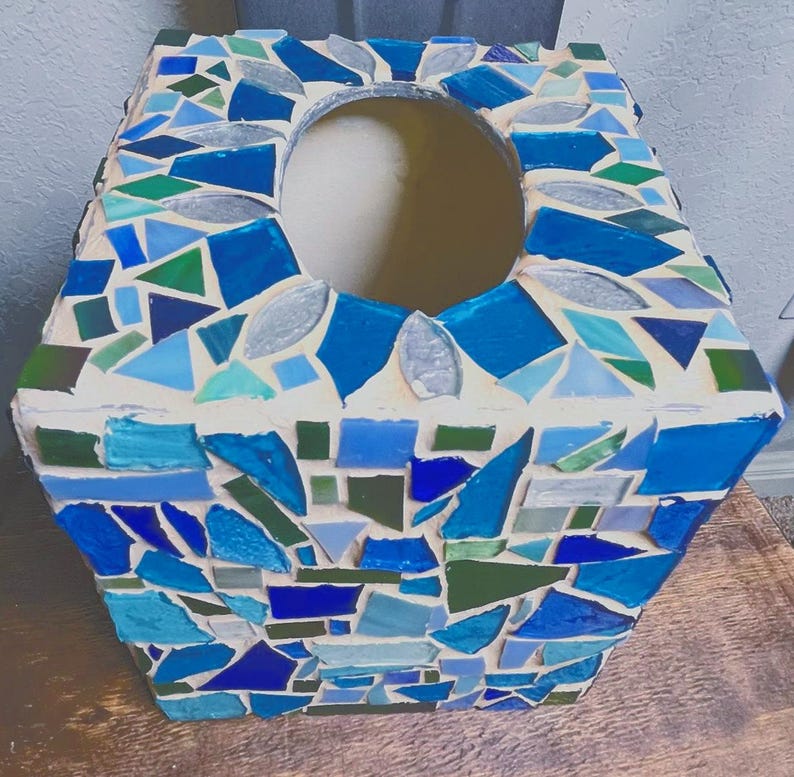 Op de afbeelding: Een vierkante tissueboxhouder met een moza&iuml;ekontwerp van diverse tinten blauw, groen en helder glas, ingebed in lichtgekleurde voegspecie. De bovenzijde is voorzien van een centrale ovale opening voor het uitgeven van tissues.