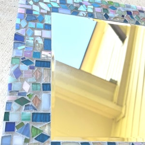 Op de afbeelding: Een vierkante spiegel met een decoratieve rand bestaande uit kleine, verschillend gevormde moza&iuml;ektegels in diverse tinten blauw, groen en iriserend wit. De tegels zijn in een dicht patroon rondom het reflecterende glasoppervlak geplaatst.