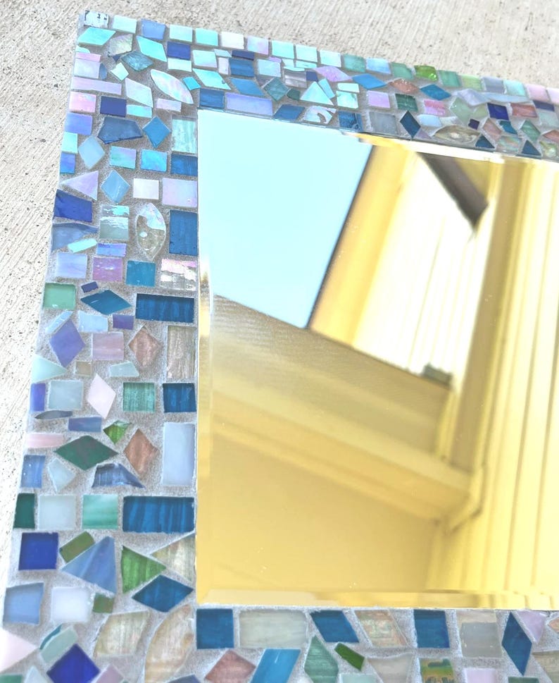 Op de afbeelding: Een vierkante spiegel met een decoratieve rand bestaande uit kleine, verschillend gevormde moza&iuml;ektegels in diverse tinten blauw, groen en iriserend wit. De tegels zijn in een dicht patroon rondom het reflecterende glasoppervlak geplaatst.