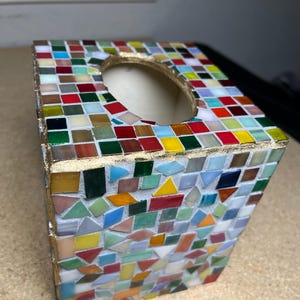 K&ouml;nnte beinhalten: Eine quadratische Taschentuchbox-Abdeckung mit einem mehrfarbigen Mosaikfliesendesign. Die Au&szlig;enseite ist mit kleinen, unregelm&auml;&szlig;ig geformten Glasfliesen in Rot-, Gelb-, Gr&uuml;n-, Blau- und Wei&szlig;t&ouml;nen verziert, die durch goldfarbene Fugen getrennt sind.