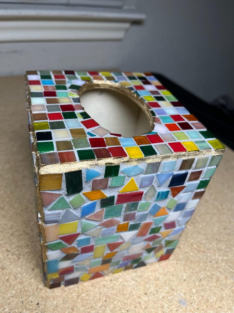 K&ouml;nnte beinhalten: Eine quadratische Taschentuchbox-Abdeckung mit einem mehrfarbigen Mosaikfliesendesign. Die Au&szlig;enseite ist mit kleinen, unregelm&auml;&szlig;ig geformten Glasfliesen in Rot-, Gelb-, Gr&uuml;n-, Blau- und Wei&szlig;t&ouml;nen verziert, die durch goldfarbene Fugen getrennt sind.
