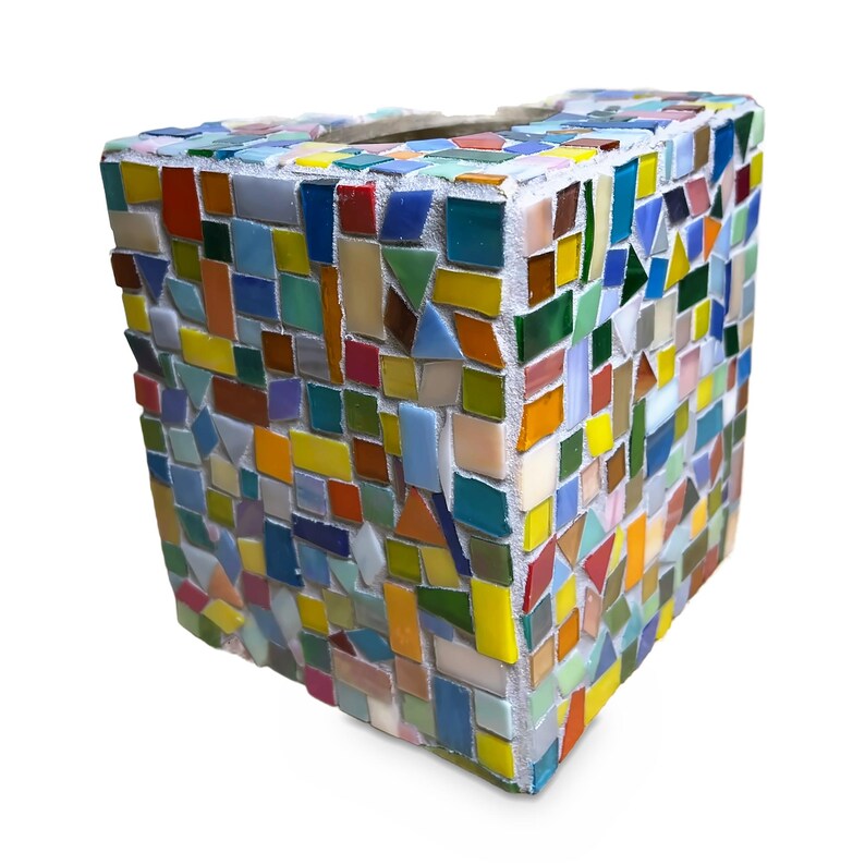 May include: A square-shaped tissue box cover decorated with a colorful mosaic pattern. The exterior is composed of numerous small, irregularly shaped glass tiles in shades of red, blue, yellow, green, orange, and white, set in white grout.