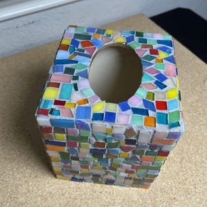 May include: A square tissue box cover decorated with a colorful mosaic pattern made of small, multi-shaped glass tiles in shades of red, blue, yellow, green, pink, and orange, featuring a central oval opening for dispensing tissues.