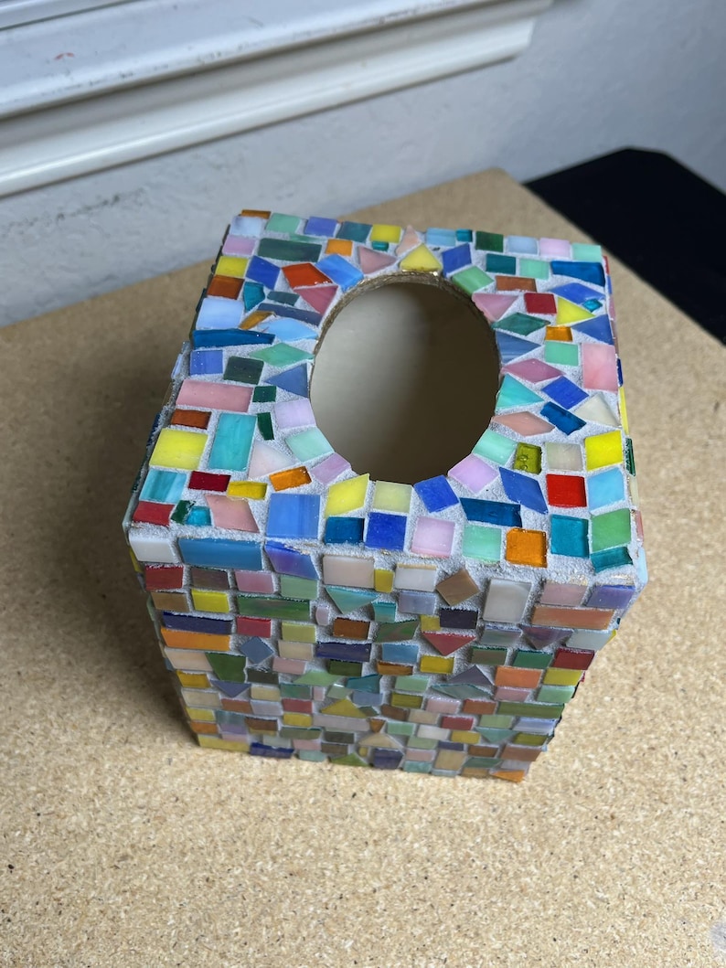 May include: A square tissue box cover decorated with a colorful mosaic pattern made of small, multi-shaped glass tiles in shades of red, blue, yellow, green, pink, and orange, featuring a central oval opening for dispensing tissues.