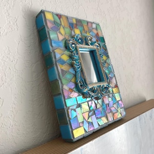 May include: A rectangular picture frame decorated with a mosaic of iridescent, blue, yellow, and pink glass tiles. Centered on the mosaic surface is a smaller, ornate, metallic blue and gold rectangular frame surrounding a reflective mirror.