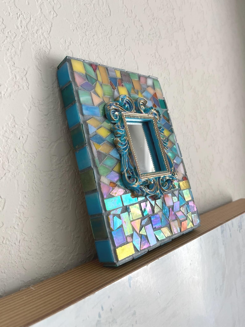 May include: A rectangular picture frame decorated with a mosaic of iridescent, blue, yellow, and pink glass tiles. Centered on the mosaic surface is a smaller, ornate, metallic blue and gold rectangular frame surrounding a reflective mirror.