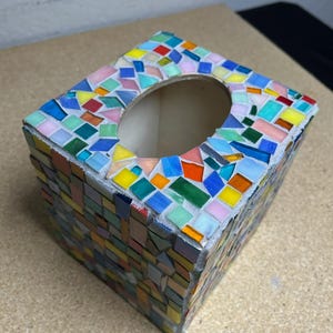May include: A square tissue box cover featuring a mosaic design made of small, multi-colored glass tiles in shades of blue, green, yellow, orange, and pink. The tiles are arranged in a random pattern with visible gray grout between each piece.