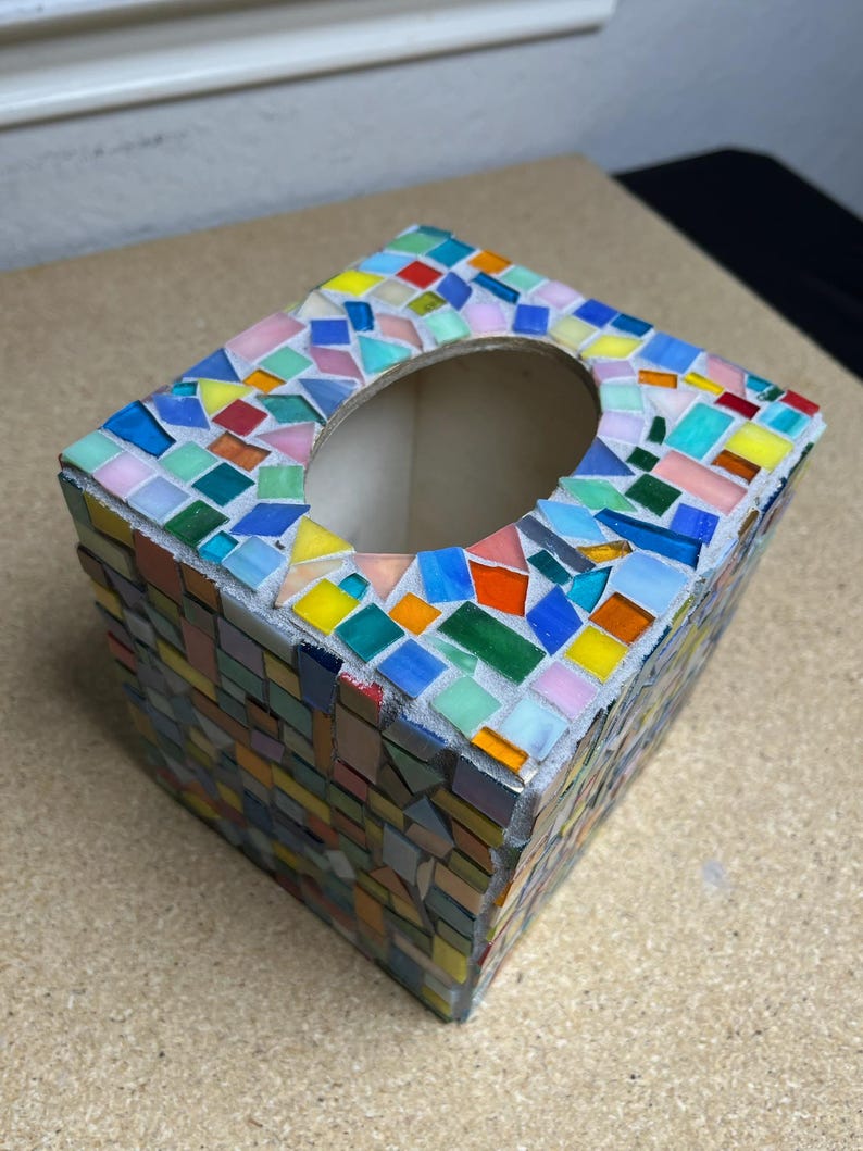 May include: A square tissue box cover featuring a mosaic design made of small, multi-colored glass tiles in shades of blue, green, yellow, orange, and pink. The tiles are arranged in a random pattern with visible gray grout between each piece.