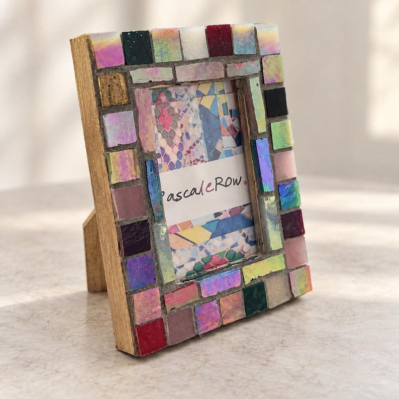 May include: A rectangular picture frame with a wooden base and a mosaic border made of small, multicolored square tiles in shades of pink, red, black, and iridescent white. The center contains a white insert with the text PascaleRow.