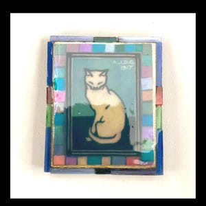 May include: A rectangular decorative tile featuring a central illustration of a seated cream-colored cat against a dark blue background. The image includes the text A.J.D.G 1917. A border of small, multicolored glass tiles surrounds the central panel.