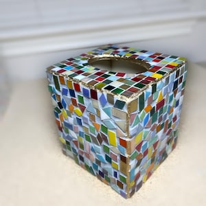 K&ouml;nnte beinhalten: Ein quadratischer Taschentuchbox-&Uuml;berzug, verziert mit einem bunten Mosaikmuster aus kleinen Glasfliesen in Rot, Blau, Gr&uuml;n, Gelb und Wei&szlig;. Die Kanten und die ovale &Ouml;ffnung an der Oberseite sind mit einer goldfarbenen Umrandung auf wei&szlig;em Fugenm&ouml;rtel versehen.