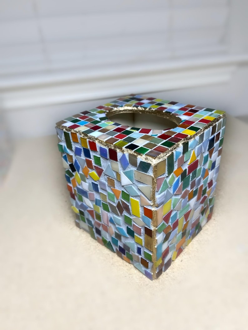 K&ouml;nnte beinhalten: Ein quadratischer Taschentuchbox-&Uuml;berzug, verziert mit einem bunten Mosaikmuster aus kleinen Glasfliesen in Rot, Blau, Gr&uuml;n, Gelb und Wei&szlig;. Die Kanten und die ovale &Ouml;ffnung an der Oberseite sind mit einer goldfarbenen Umrandung auf wei&szlig;em Fugenm&ouml;rtel versehen.