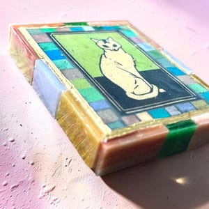 May include: A rectangular resin block featuring a central illustration of a seated cat on a green and black background. The border consists of a mosaic-style pattern of small, multicolored translucent squares in shades of blue, green, and gold.