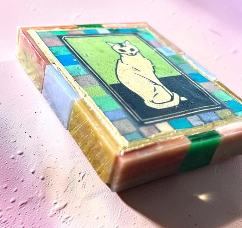 May include: A rectangular resin block featuring a central illustration of a seated cat on a green and black background. The border consists of a mosaic-style pattern of small, multicolored translucent squares in shades of blue, green, and gold.